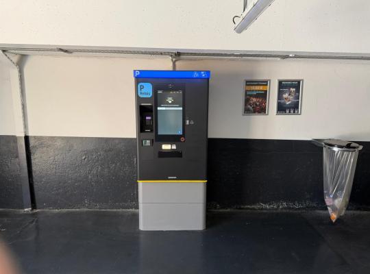 Caisse automatique de paiement Skidata installée contre un mur avec affichage des tarifs à proximité.