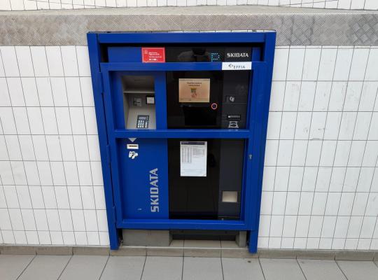 Caisse automatique de paiement Skidata encastrée dans un cadre bleu sur un mur carrelé blanc.