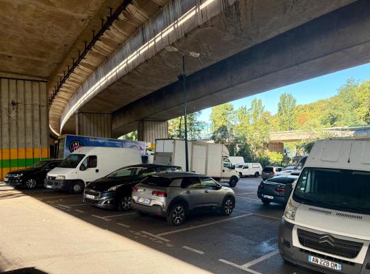 Véhicules et utilitaires garés sur des emplacements tracés sous les piliers d'un viaduc.