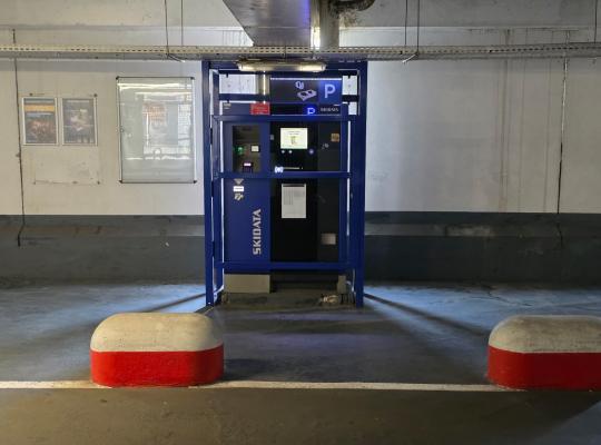 Caisse automatique Skidata protégée par une structure métallique bleue à l'intérieur du parking couvert.