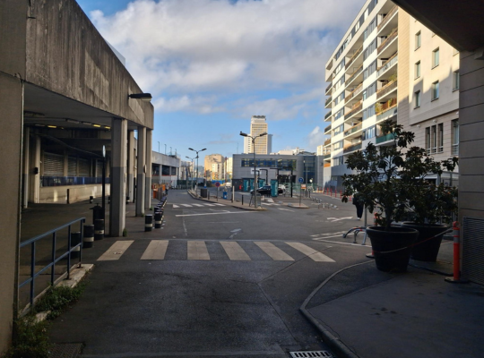Perspective depuis un passage couvert donnant sur une rue extérieure avec des immeubles et des bacs de fleurs.