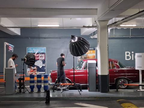 Équipe de tournage vidéo installée avec du matériel d'éclairage professionnel devant une voiture ancienne dans un parking couvert.